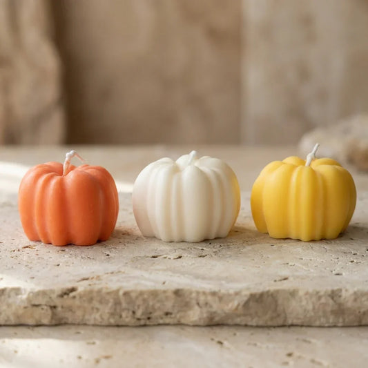 Three pumpkin-shaped candles in orange, white, and yellow on a stone surface.