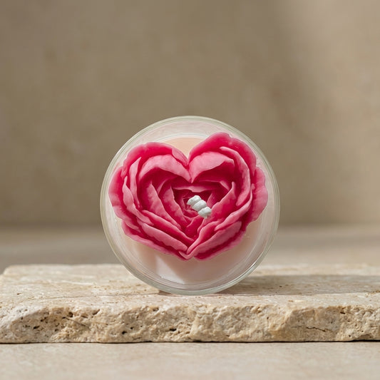 Close-up of a clear glass candle jar featuring an intricately sculpted, vibrant pink floral heart resting on top of natural white soy wax.