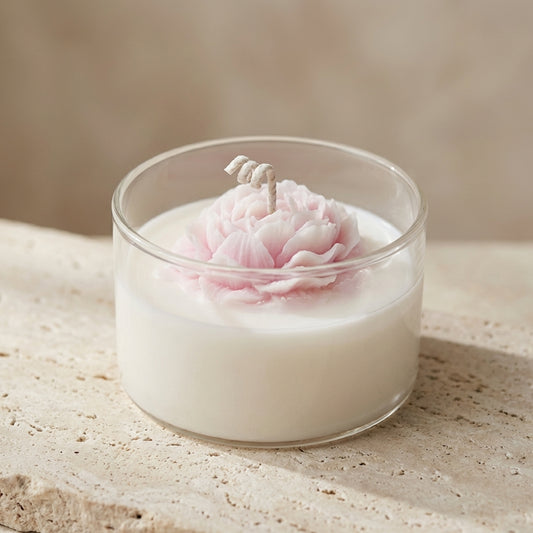 Close-up of a clear glass candle jar featuring an intricately sculpted, pale pink wax peony blossom resting on top of natural white soy wax.