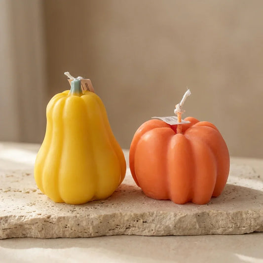 Two pumpkin-shaped candles, one yellow and one orange, on a stone surface with a neutral background.