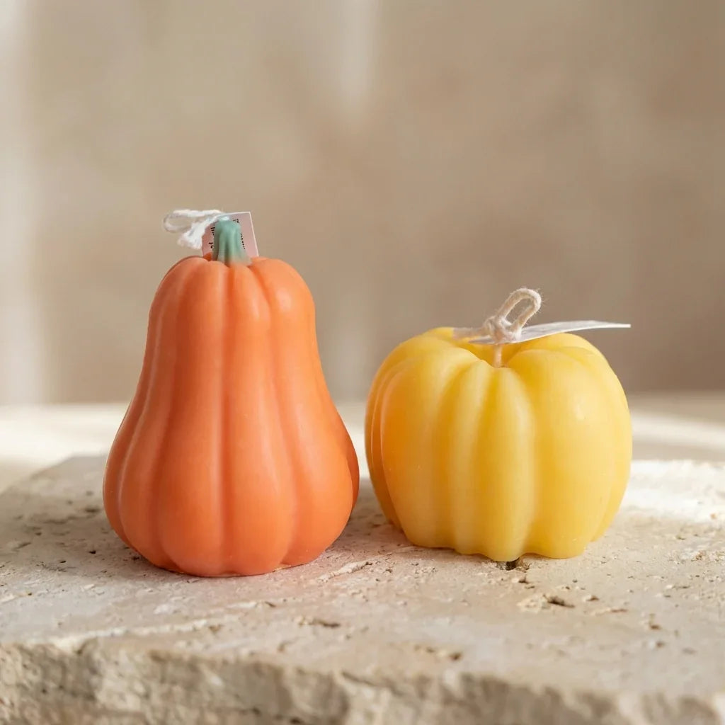 Two pumpkin-shaped candles, one orange and one yellow, on a stone surface with a neutral background.