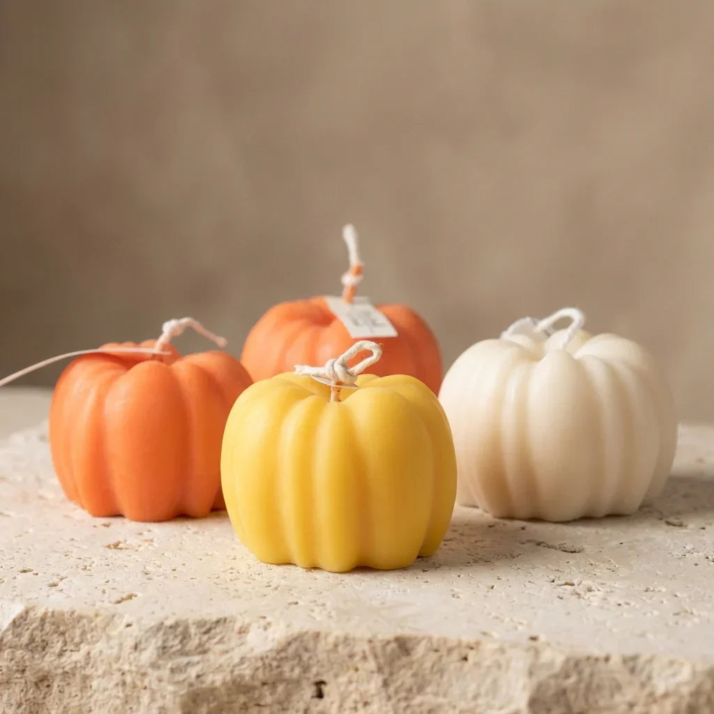Four pumpkin-shaped candles in orange, yellow, and white on a stone surface with a neutral background.